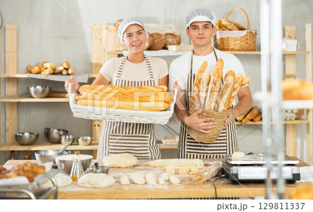Two smiling young bakers holding freshly baked goods in bakery Two smiling young bakers holding freshly baked goods in bakery 129811337