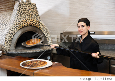 Female chef in black uniform prepares pizza and pulls it out of oven in kitchen of pizzeria 129811368
