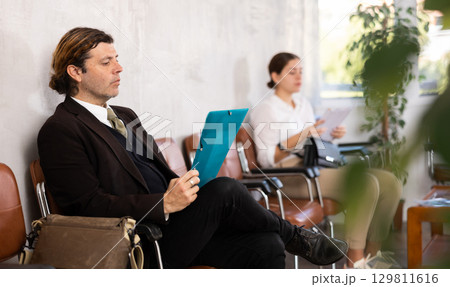 Adult man in suit with folder of documents sits on chair Adult man in suit with folder of documents sits on chair 129811616