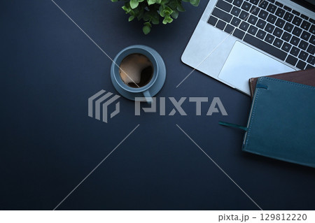 Flat lay workspace with laptop, coffee cup, notebook, and plant on a dark background 129812220