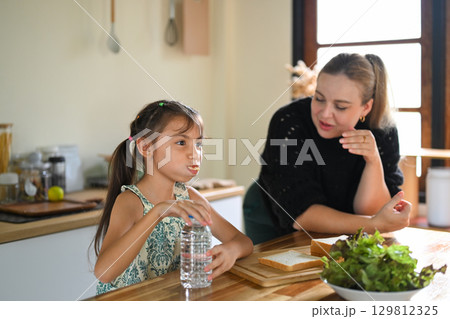 Young girl drinking water and eating at the kitchen table while her mother watches with a smile Young girl drinking water and eating at the kitchen table while her mother watches with a smile 129812325