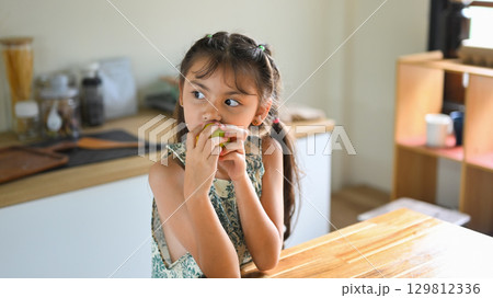 Cute little girl eating a fresh green apple in the kitchen, enjoying a healthy snack Cute little girl eating a fresh green apple in the kitchen, enjoying a healthy snack 129812336