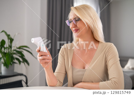 Cheerful blonde middle-aged woman in beige outfit looking happily at thermometer while feeling better in modern apartment living room 129815749