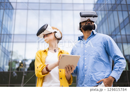 A diverse couple uses virtual reality headsets outdoors, experiencing augmented reality with a tablet in front of a modern building. 129815784