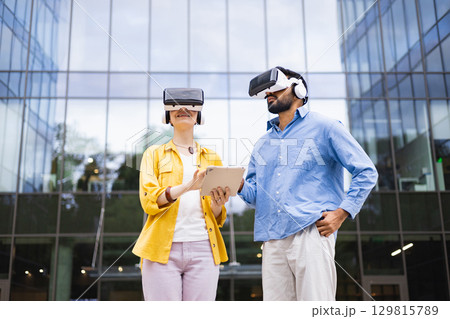 Two people wearing VR headsets are standing in front of a modern building, likely exploring virtual reality with a tablet. Two people wearing VR headsets are standing in front of a modern building, likely exploring virtual reality with a tablet. 129815789