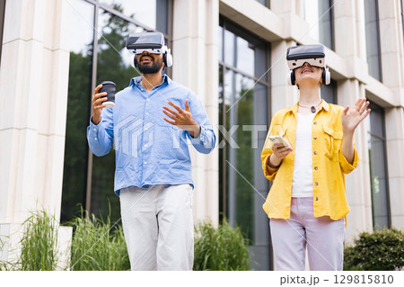 A diverse couple experiences virtual reality outdoors. They wear VR headsets and interact with their surroundings. The man holds a coffee cup. 129815810