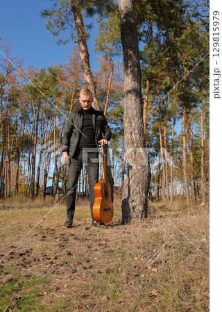 Blonde man playing string guitar outdoors in autumn forest. Concept of sound therapy, mental health and wellness rituals. Calmness tranquility audio-sensory practices. Aura farming energy 129815979