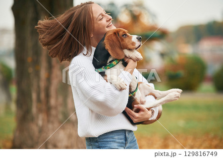Standing against tree, holding animal in hands. Young woman is with her dog in the park 129816749