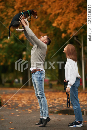 Man is holding animal high up in the hands. Lovely couple are with their cute dog outdoors Man is holding animal high up in the hands. Lovely couple are with their cute dog outdoors 129816819