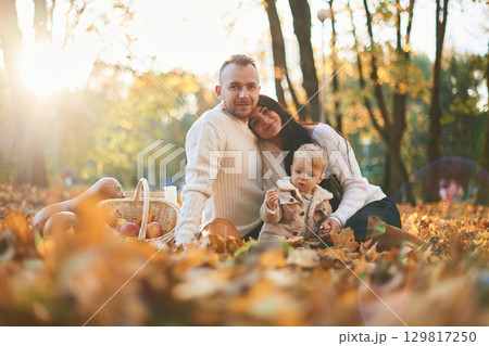 Two parents are sitting with cute little boy in the autumn park on the fallen leaves and butternut squash Two parents are sitting with cute little boy in the autumn park on the fallen leaves and butternut squash 129817250