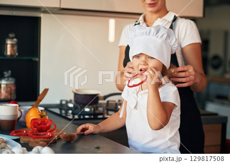 Girl is holding pepper. Mother with her daughter are preparing food on the kitchen Girl is holding pepper. Mother with her daughter are preparing food on the kitchen 129817508