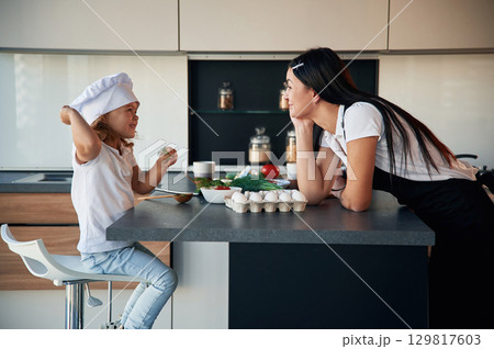 Girl is in the chief hat. Mother with her daughter are preparing food on the kitchen Girl is in the chief hat. Mother with her daughter are preparing food on the kitchen 129817603