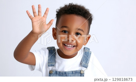 Happy african child smiling and waving in denim overalls against white background. 129818673
