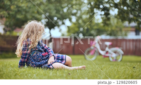 Blond child sitting on verdant lawn, gazing at bicycle while enjoying carefree family moment in sunlit backyard 129820605