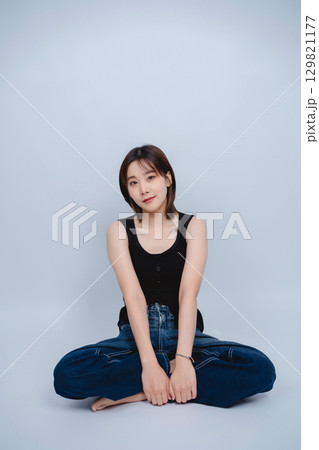 Young woman with short hair wearing a black tank top and denim jeans, sitting on the floor with a calm expression against a plain gray background 129821177