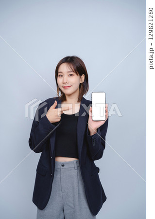 Confident Young Woman in Business Attire Pointing at Smartphone with Blank Screen, Showcasing Modern Technology and Professional Style in Minimalist Background Confident Young Woman in Business Attire Pointing at Smartphone with Blank Screen, Showcasing Modern Technology and Professional Style in Minimalist Background 129821280