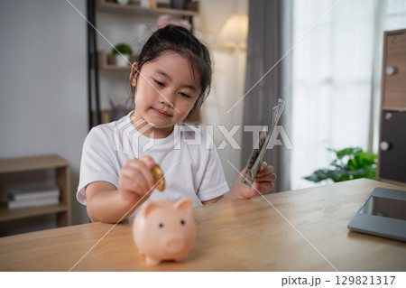 Young girl saving coin in piggy bank at home, financial literacy, childhood savings habit, teaching children about money management 129821317