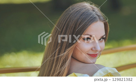 Beautiful young woman with long brown hair, wearing a yellow top, turning and smiling over her shoulder in a park Beautiful young woman with long brown hair, wearing a yellow top, turning and smiling over her shoulder in a park 129821382