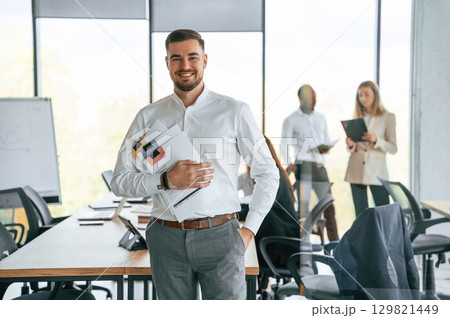 Cheerful man is with documents in hands, standing. Group of office workers are together indoors Cheerful man is with documents in hands, standing. Group of office workers are together indoors 129821449