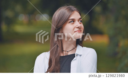 Portrait of a cheerful young woman with long brown hair, wearing a white shirt, smiling and looking up in a park Portrait of a cheerful young woman with long brown hair, wearing a white shirt, smiling and looking up in a park 129821980