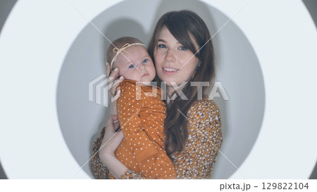 Mother smiling while holding her baby daughter, enveloped in a warm circle of light, capturing a beautiful moment of love and togetherness 129822104