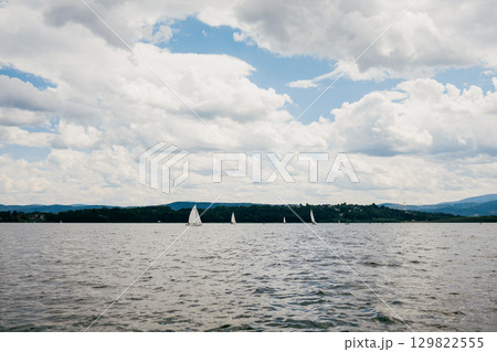 Sailboats gliding across tranquil waters of lake, enjoying beautiful day under cloudy sky, with majestic mountains rising in background, create picturesque scene. Sailboats, lake, mountainous, sky Sailboats gliding across tranquil waters of lake, enjoying beautiful day under cloudy sky, with majestic mountains rising in background, create picturesque scene. Sailboats, lake, mountainous, sky 129822555