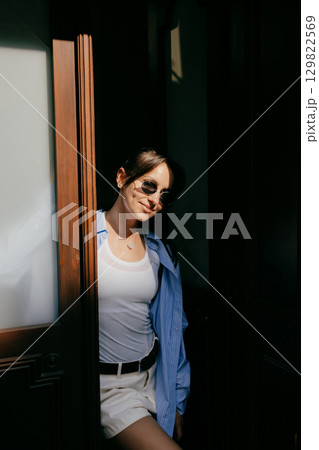 Fashionable young woman with sunglasses standing in doorway, enjoying sunlight filtering through, creating contrast between light and shadow. Stylish woman in sunglasses leaning against doorway Fashionable young woman with sunglasses standing in doorway, enjoying sunlight filtering through, creating contrast between light and shadow. Stylish woman in sunglasses leaning against doorway 129822569