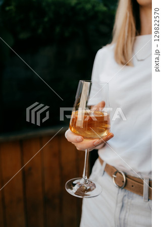 Woman is holding glass of white wine, enjoying refreshing drink on summer day against blurred wooden background. Young adult woman holding glass of white wine, relaxing during sunny summer day Woman is holding glass of white wine, enjoying refreshing drink on summer day against blurred wooden background. Young adult woman holding glass of white wine, relaxing during sunny summer day 129822570