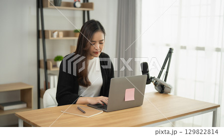 Young woman working on laptop in a modern home office with microphone and bookshelf, bright lighting and organized workspace 129822814
