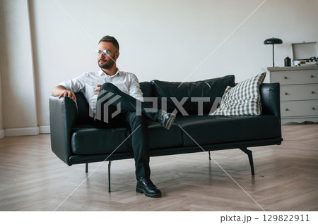 Sitting on the sofa. Elegant man in business clothes is indoors with glass of water 129822911