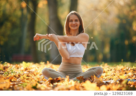 Smiling, warm up exercises for hands. Young beautiful woman is doing yoga in the autumn park 129823169