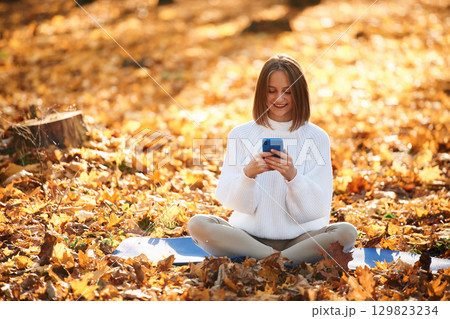 Woman is sitting in the autumn park or forest with smartphone in hands Woman is sitting in the autumn park or forest with smartphone in hands 129823234