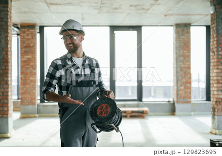 Standing and holding wires in hands. Construction worker in uniform in empty unfinished room Standing and holding wires in hands. Construction worker in uniform in empty unfinished room 129823695