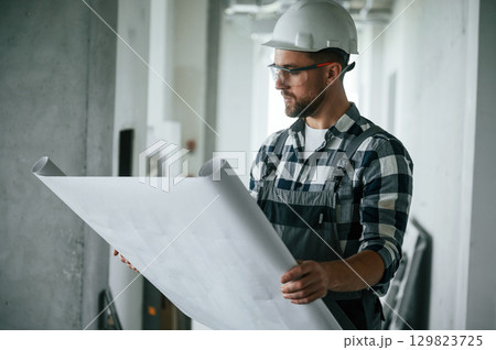 Project on the paper, holding in hands. Construction worker in uniform in empty unfinished room 129823725