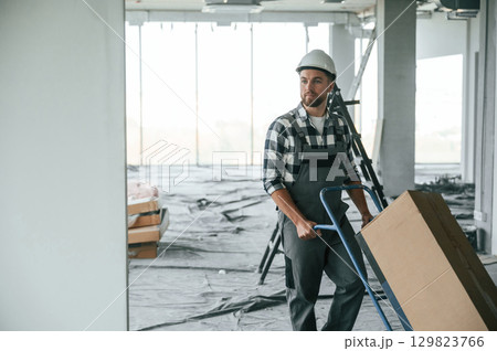 With large box, transporting the material. Construction worker in uniform in empty unfinished room With large box, transporting the material. Construction worker in uniform in empty unfinished room 129823766