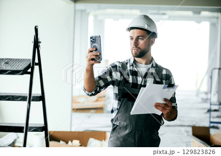 Documents in hands. In white hard hat. Repairman in uniform is in the unfinished building 129823826