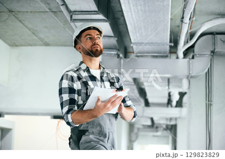 Documents in hands. In white hard hat. Repairman in uniform is in the unfinished building Documents in hands. In white hard hat. Repairman in uniform is in the unfinished building 129823829