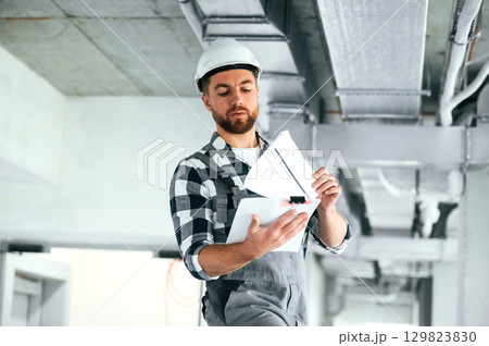Documents in hands. In white hard hat. Repairman in uniform is in the unfinished building 129823830