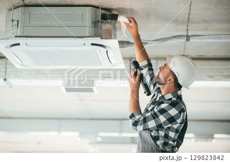 Ventilation system high up on the ceiling, fixing. Repairman in uniform is in the unfinished building 129823842
