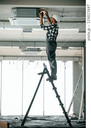 Standing on the ladder. Repairman in uniform is in the unfinished building 129823847
