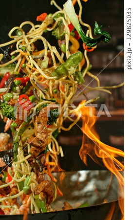 Freeze Motion of Wok Pan with Flying Ingredients in the Air and Fire Flames. Kitchen Interior on Background. Vertical Aspect Ratio. Freeze Motion of Wok Pan with Flying Ingredients in the Air and Fire Flames. Kitchen Interior on Background. Vertical Aspect Ratio. 129825035