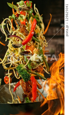 Freeze Motion of Wok Pan with Flying Ingredients in the Air and Fire Flames. Kitchen Interior on Background. Vertical Aspect Ratio. Freeze Motion of Wok Pan with Flying Ingredients in the Air and Fire Flames. Kitchen Interior on Background. Vertical Aspect Ratio. 129825041