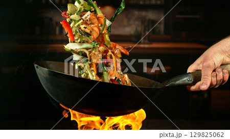 Freeze Motion of Wok Pan with Flying Ingredients in the Air and Fire Flames. Kitchen Interior on Background. Freeze Motion of Wok Pan with Flying Ingredients in the Air and Fire Flames. Kitchen Interior on Background. 129825062