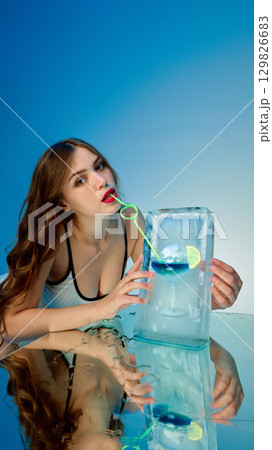 Woman sipping through straw from cocktail frozen in ice with playful charm 129826683