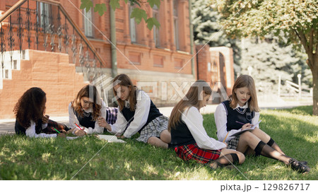 Schoolgirls studying together on green lawn with thoughtful mood 129826717