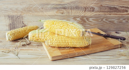Fresh corn on the cob on a cutting board with a knife on a wooden background, fresh vegetables, healthy food. Fresh corn on the cob on a cutting board with a knife on a wooden background, fresh vegetables, healthy food. 129827256