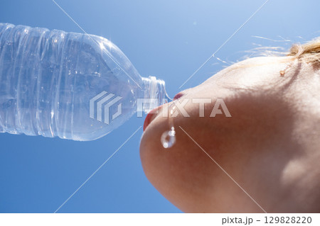Young woman enjoying refreshing water from a plastic bottle under a bright blue summer sky, quenching her thirst on a hot day 129828220