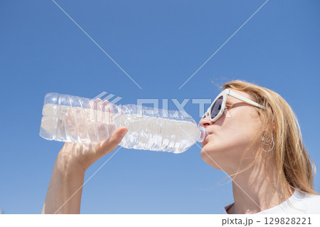 Young woman enjoying a refreshing drink of water from a plastic bottle under a bright blue sky on a hot summer day Young woman enjoying a refreshing drink of water from a plastic bottle under a bright blue sky on a hot summer day 129828221