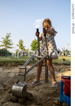 Little girl digging in the sand with a playground excavator, enjoying outdoor playtime on a sunny day 129828247