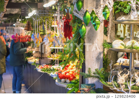 Christmas ornaments at festive market stall. Christmas market stall filled with shiny green and gold ornaments, pine decor, glowing lights. Concept handmade decorations and festive market charm 129828400